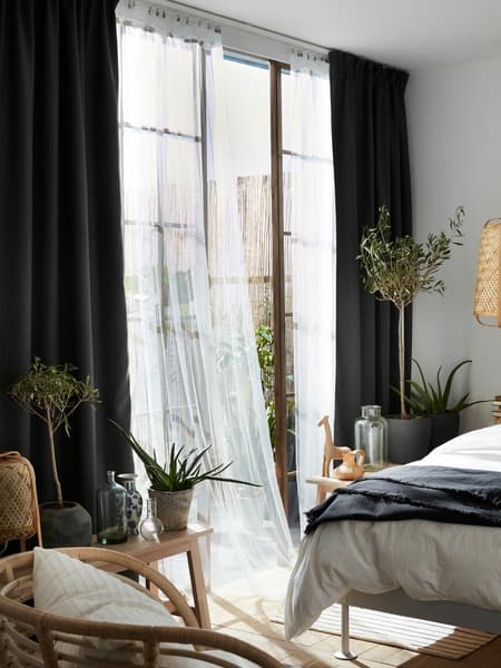 A white LILL net curtain blows in the wind from an open window in a bedroom on a sunny day, almost touching the bed.