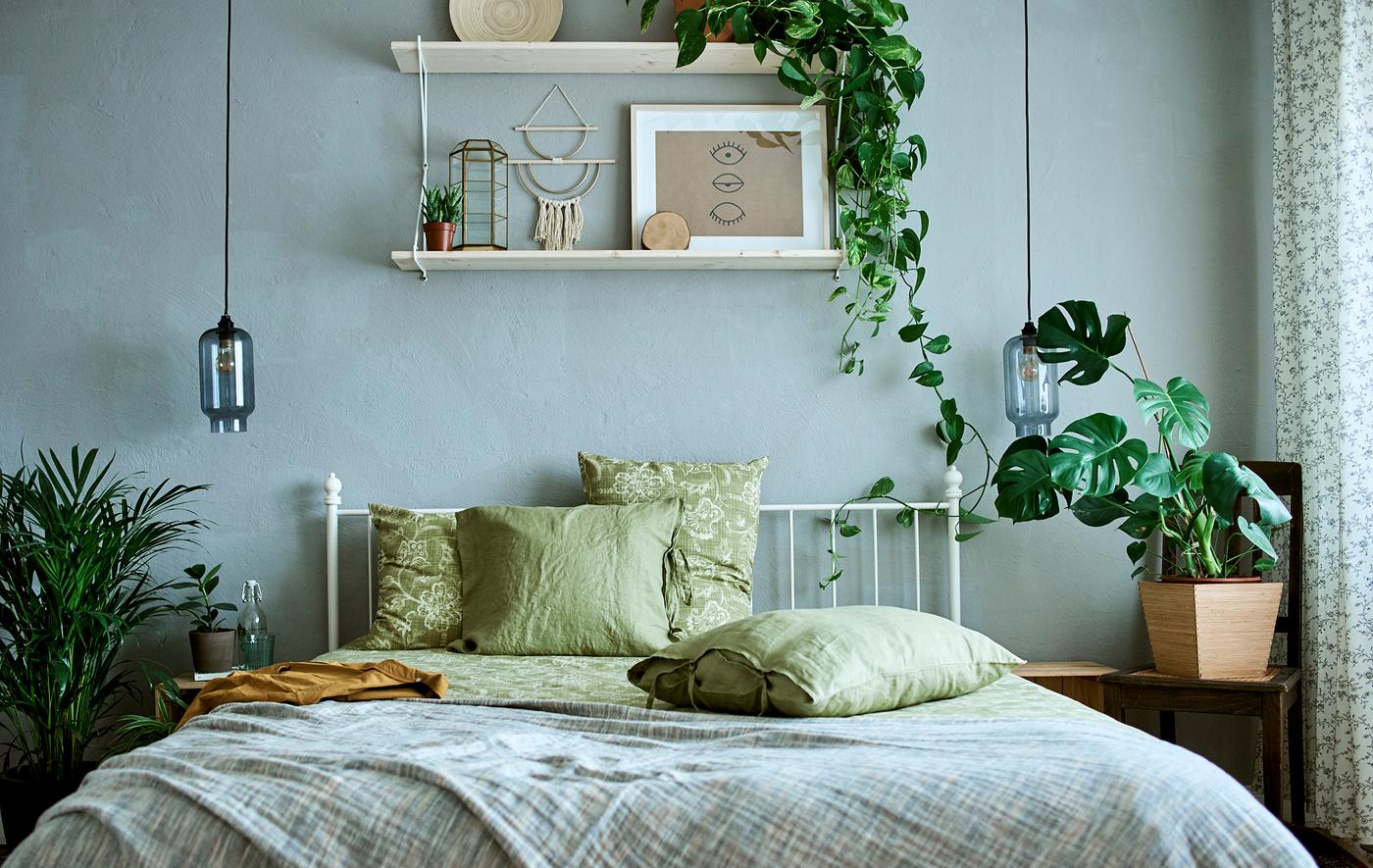 A white, metal framed bed with green bedding, plain and patterned in a room decorated with plants and art.