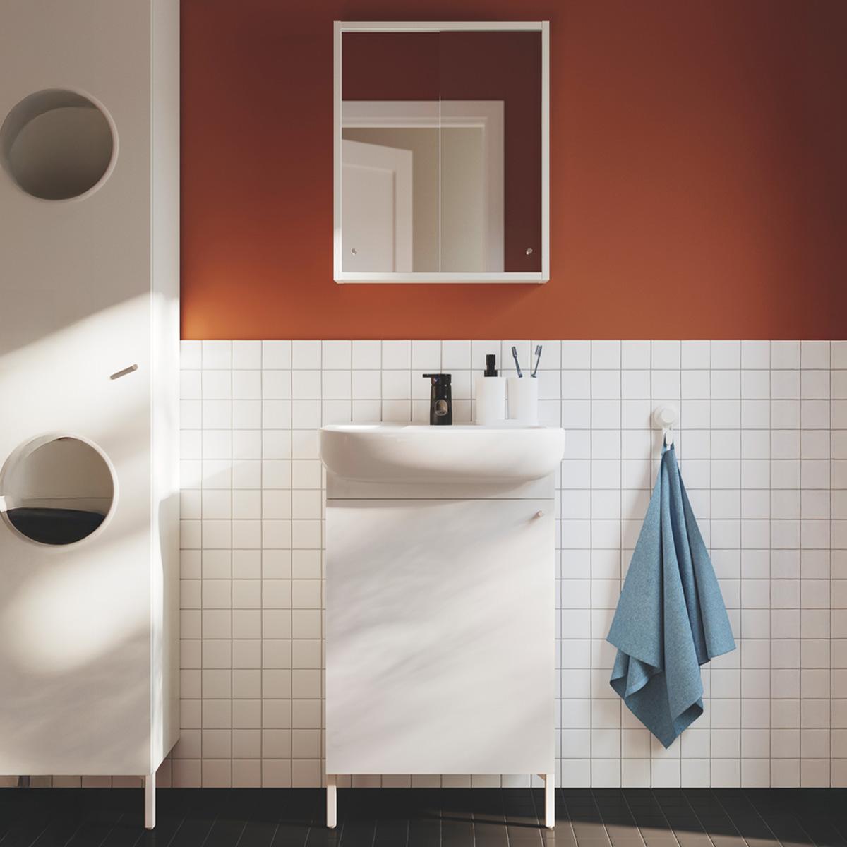 A white NYSJÖN wash-stand with door with a white BJÖRKÅN wash-basin with tap leaned against the white square tile and a white framed mirror mounted on the orange/red wall. A white NYSJÖN
laundry cabinet is seen on the left side of the bathroom, while a light blue towel is hung on the right of the wash-stand.