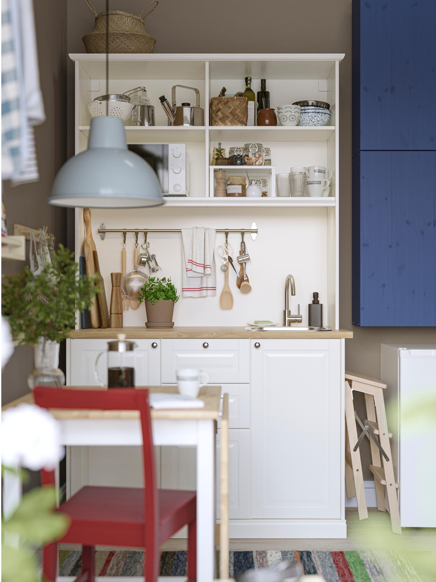 A white ÖNNERUP kitchenette has blue-painted IVAR cabinets mounted next to it and a small table with a red chair in front.