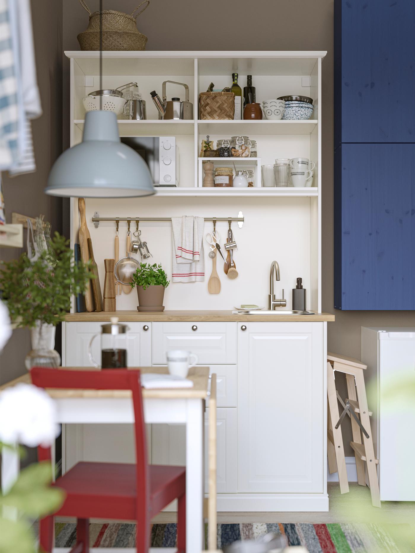 A white ÖNNERUP kitchenette has blue-painted IVAR cabinets mounted next to it and a small table with a red chair in front.