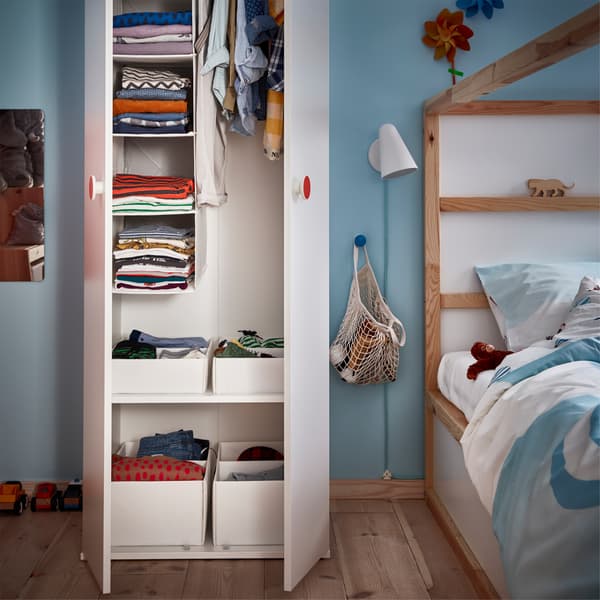 A white open closet with organised children's clothes, a bed in white and pine and a white wall lamp.