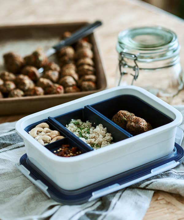 A white plastic food tub with blue inserts, each contains different food, on a linen tea towel beside a tray of veggie balls.
