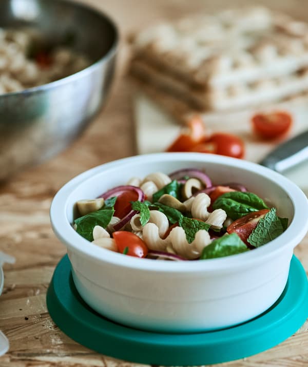 A white plastic round tub containing tomato, red onion and green olive pasta salad, on top of a blue lid on a wood table top.