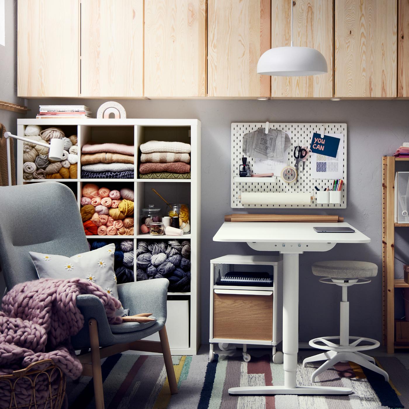 A white shelving unit with yarn, a light green armchair, a white desk with sit/stand support, pine cabinets, and a pendant.