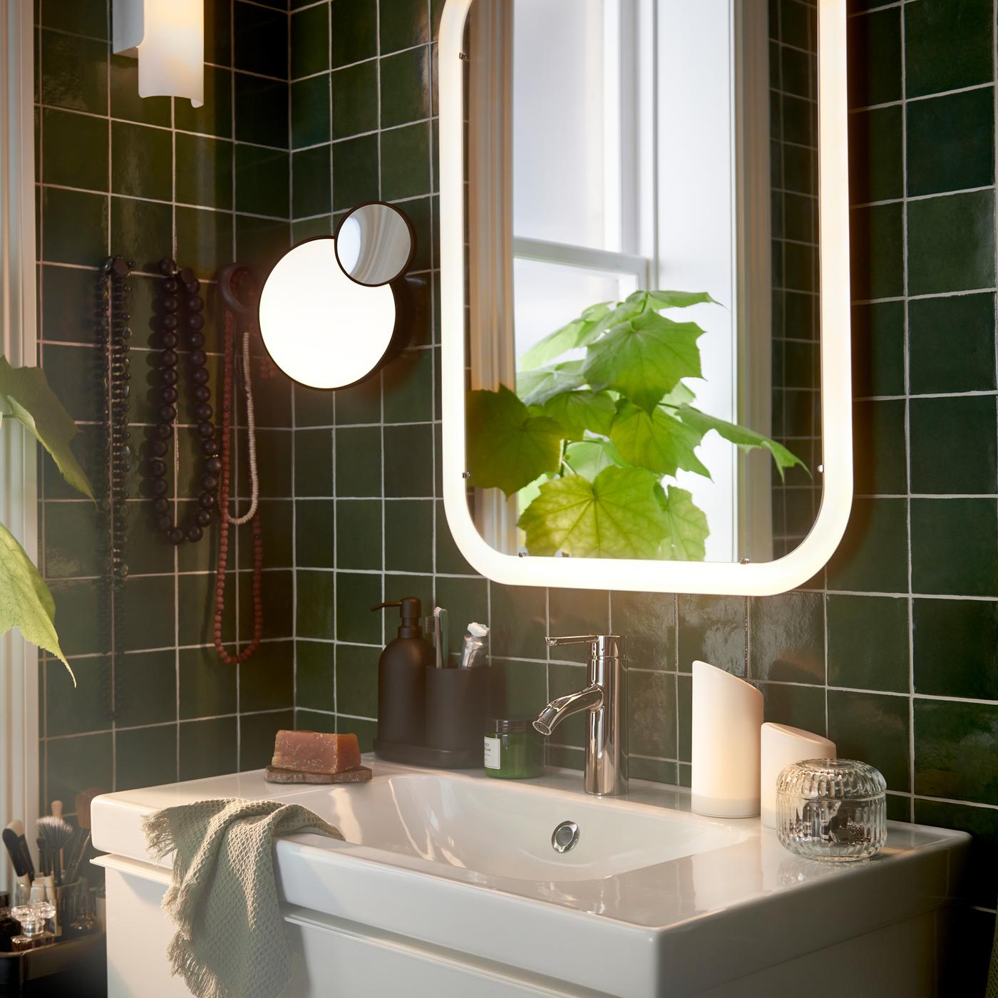 A white sink and a STORJORM mirror with integrated lighting placed in a small bathroom with dark green tiles.
