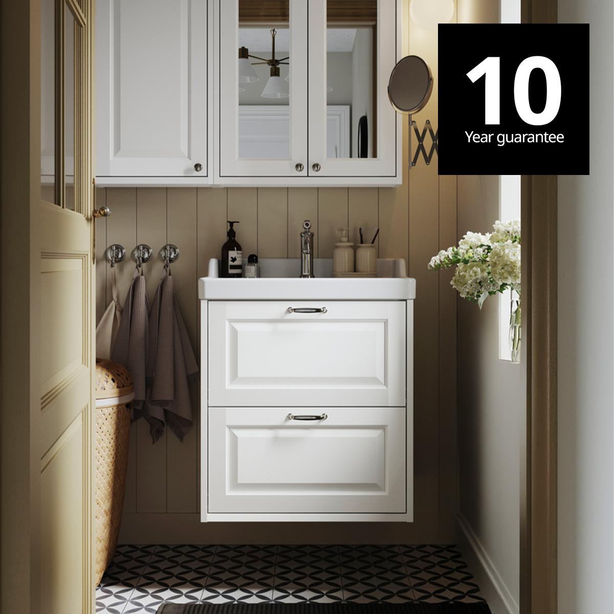 A white TÄNNFORSEN wash-stand with drawers and a matching mirror cabinet are placed in a bathroom with light brown walls. 10-year guarantee logo on top right corner.