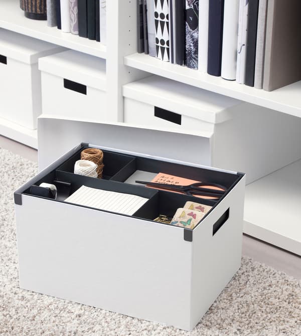A white TJENA storage box filled with stationery, placed on a light grey carpet, with more boxes in the background.