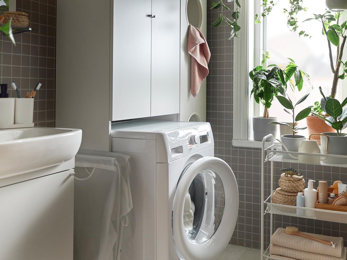 A white washing machine in a bathroom, placed inside ENHET laundry solution. Plants in the window and a cart next to it.