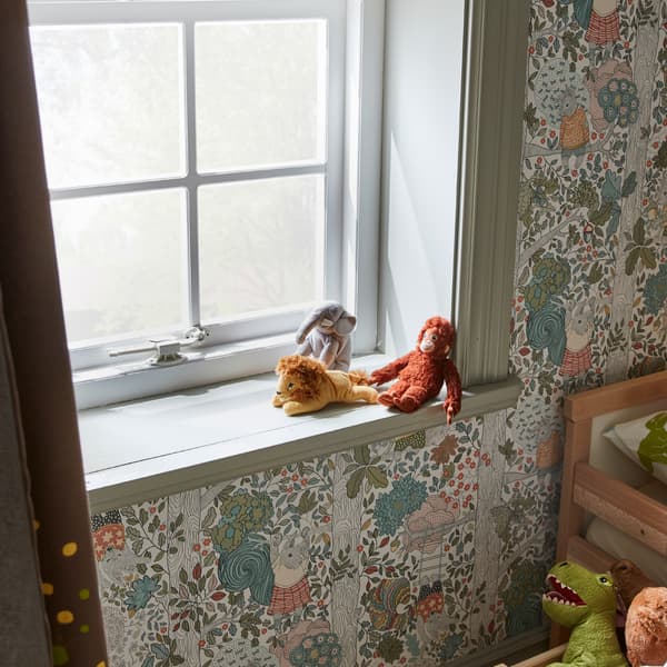 A window with a white window catch, a green window sill with three soft toys and a forest-patterned wallpaper on the walls.
