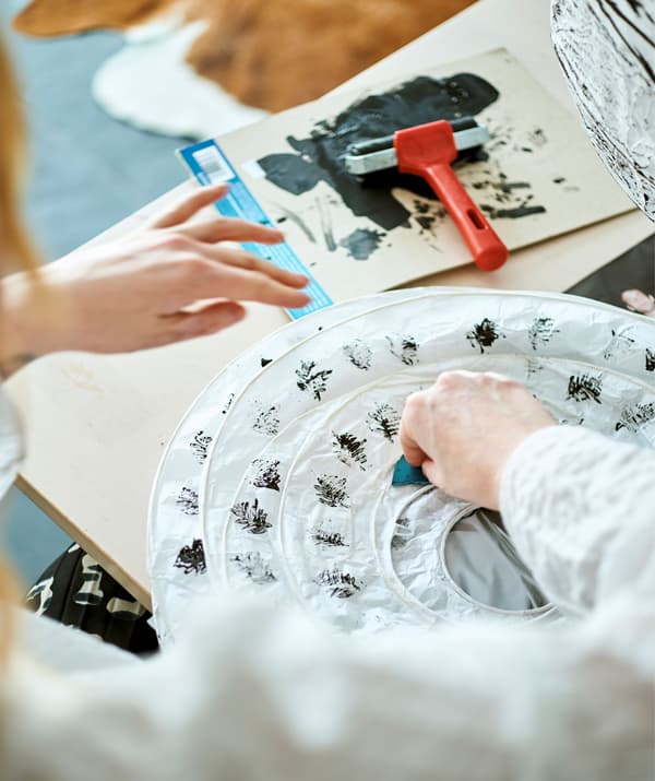 A woman adds a pattern of fern leaves to a folded white paper lantern by pressing on an ink stamp dipped in black paint.