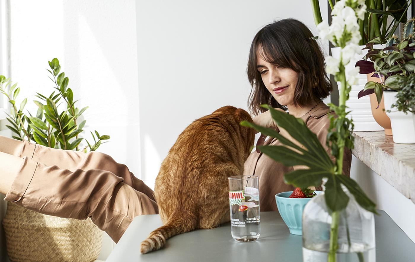 A woman and cat sitting at a table on a white balcony with lots of pot plants.