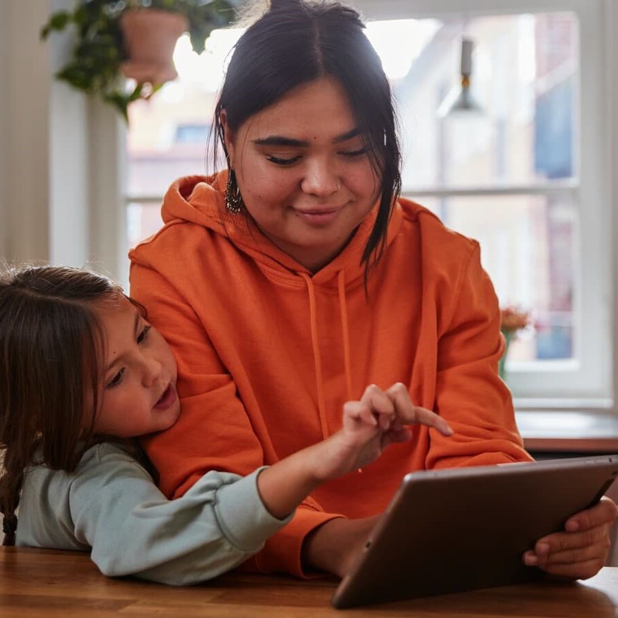 A woman and her child are sitting in the kitchen and looking at a tablet together.