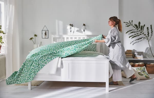 A woman dressed in a white flowing linen robe drapes a leafy green sheet over a HEMNES bed.