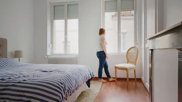 A woman dressed in jeans and a t-shirt standing looking out her bedroom window.