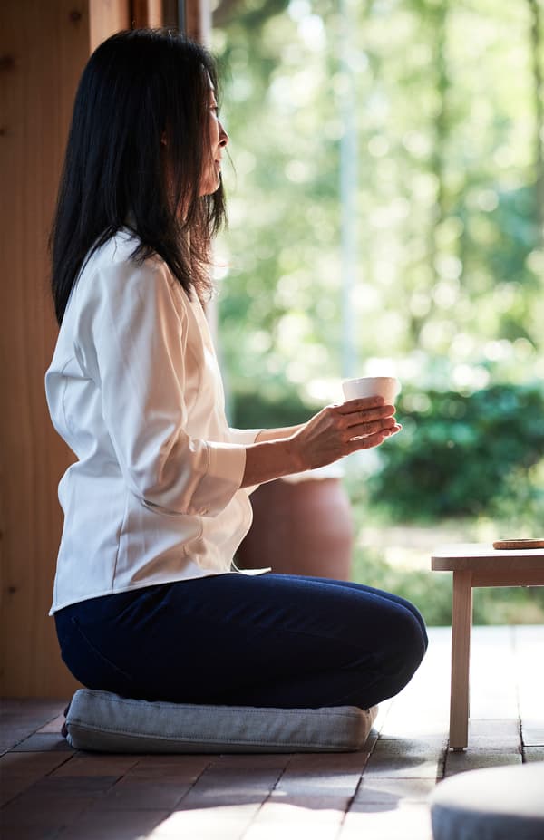 A woman holding a mug in both hands sits on an OMTÄNKSAM chair cushion placed on the floor.