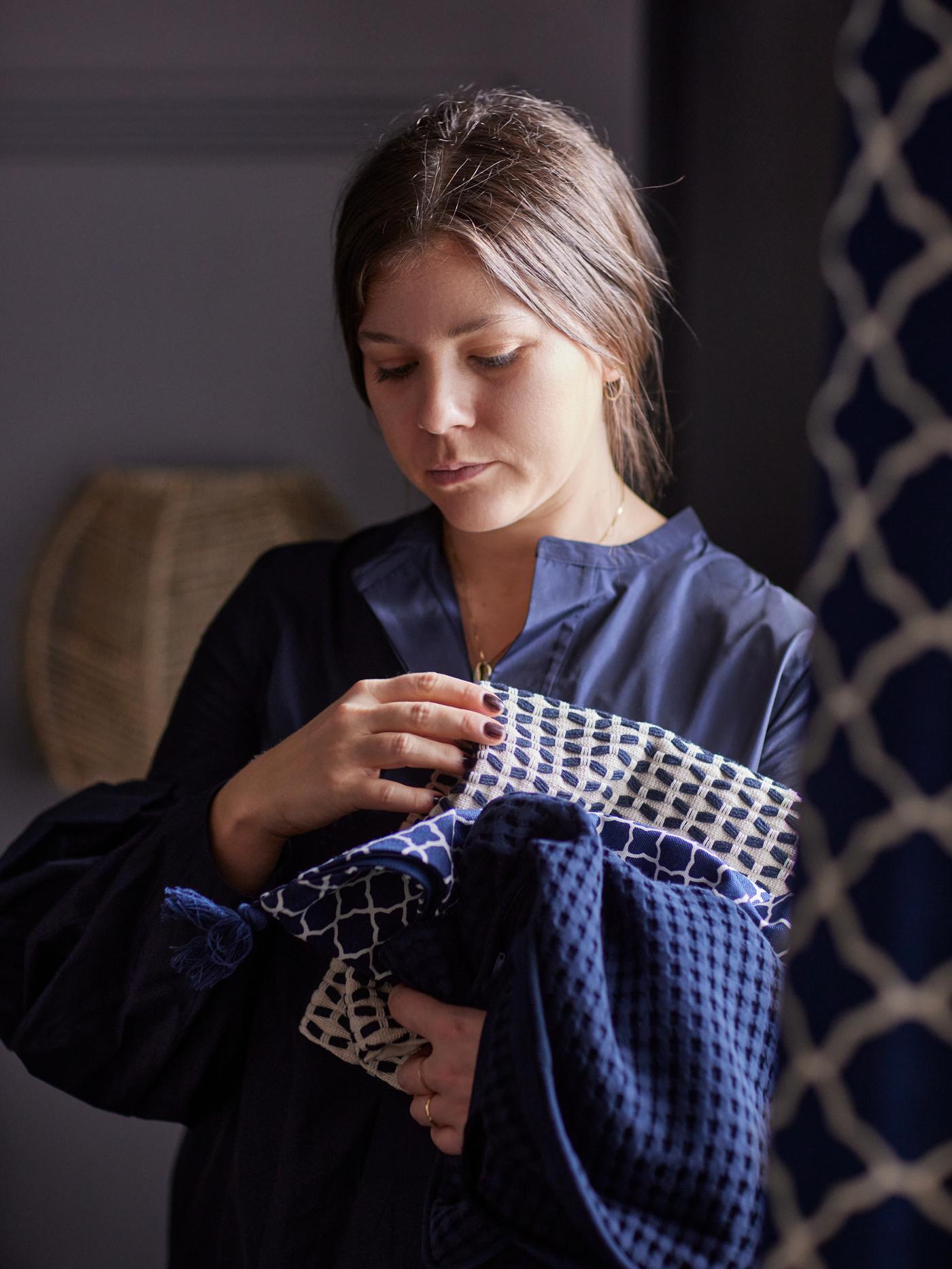 A woman holding folded bedroom textiles in different blue tones and patterns in her arms.