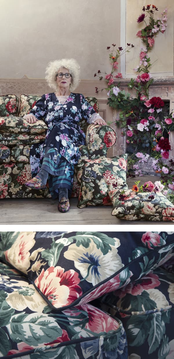A woman in a floral dress on a floral sofa next to a fireplace dressed with flowers.