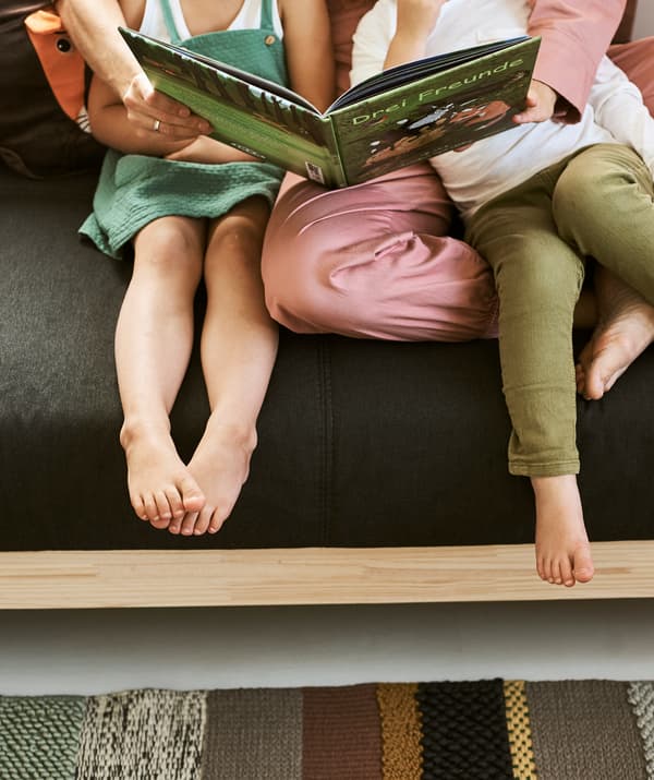 A woman in a pink jumpsuit sits on a grey and wood sofa with her arms around a boy and girl, reading a children’s book.