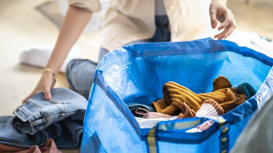 A woman in a shirt with sleeves rolled up folding clothes and placing them in a big blue IKEA bag on the floor.