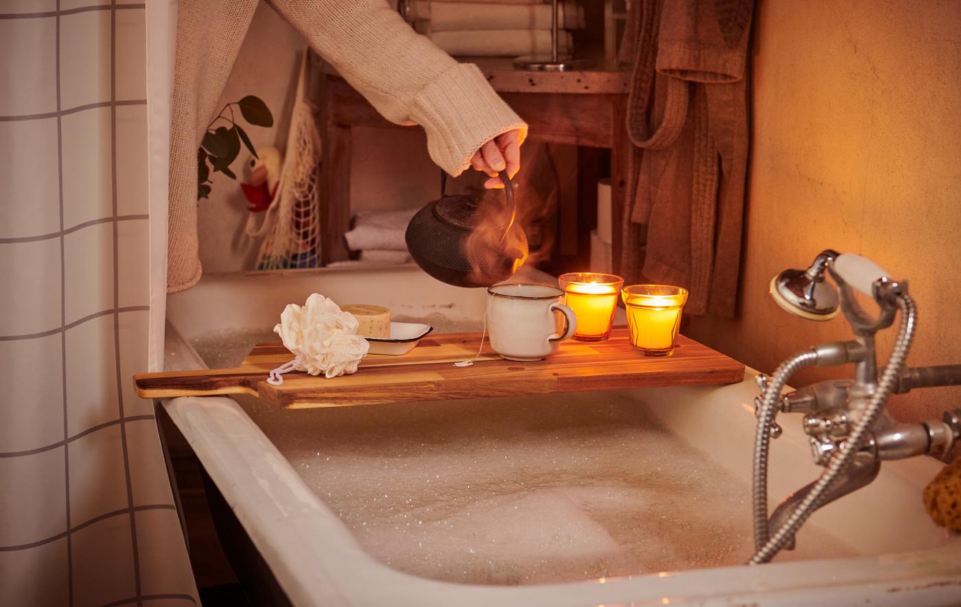 A woman in a sweater preparing for a spa day pours tea into a mug on a cutting board balanced across a bathtub.