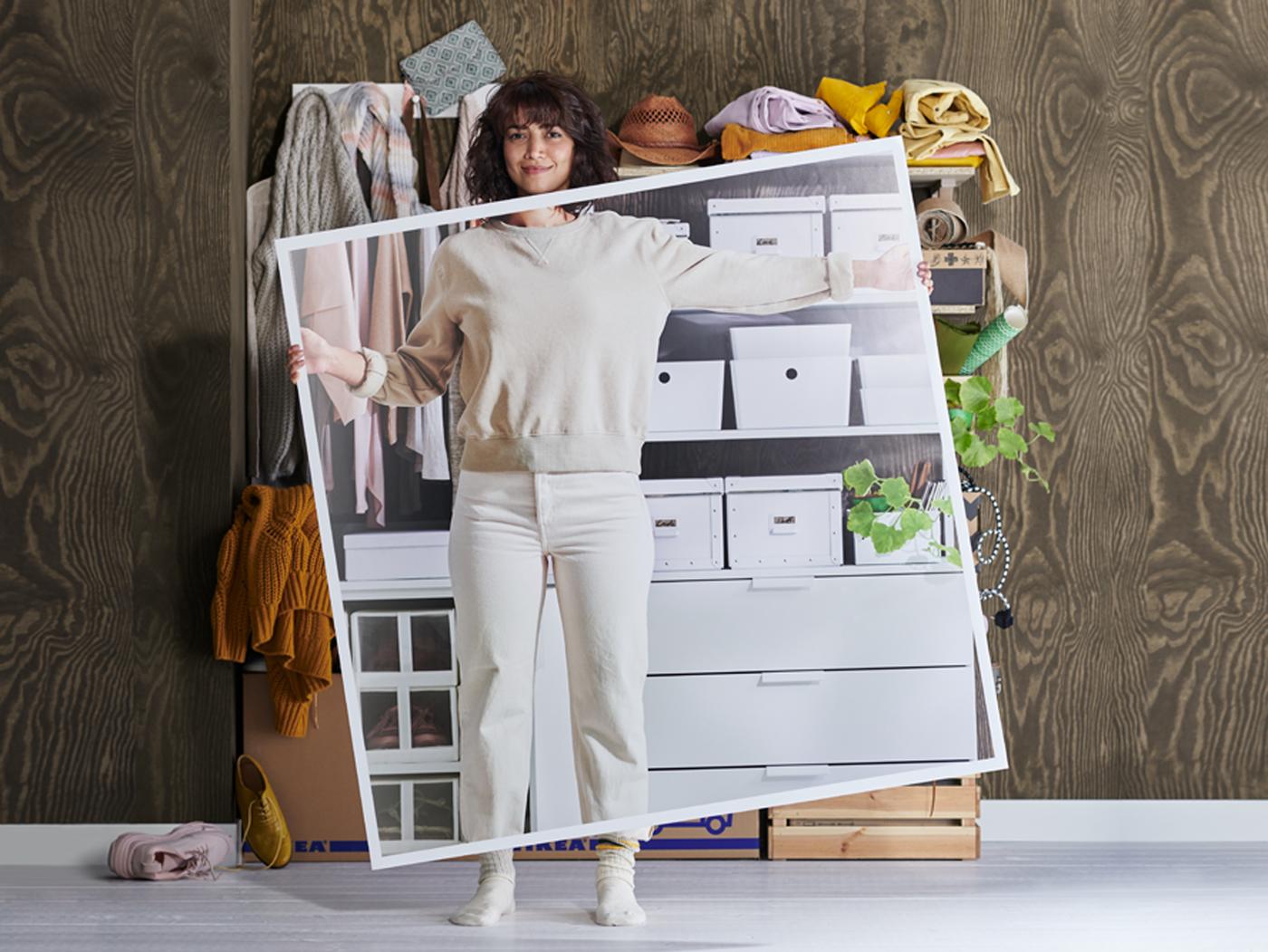 A woman in white clothes stands in front of messy shelves holding a large picture of the shelves when they've been tidied up.