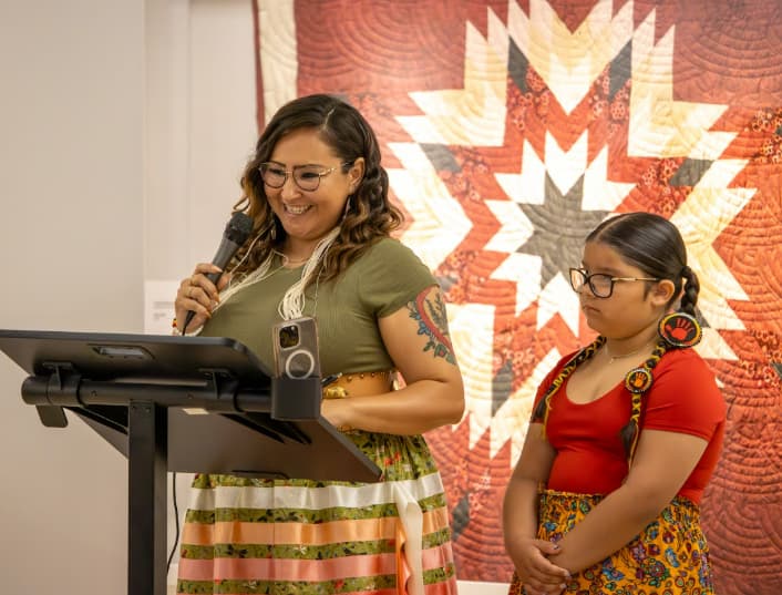 A woman is holding a microphone and is standing at a podium. A young girl stands beside her listening to her speak. 