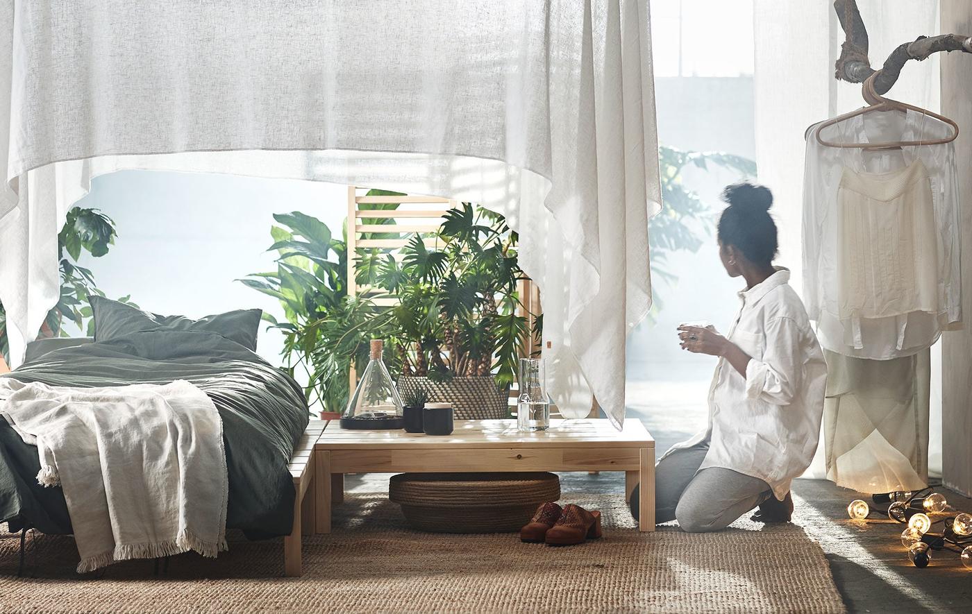 A woman kneeling next to a low table with plants next to a single bed.