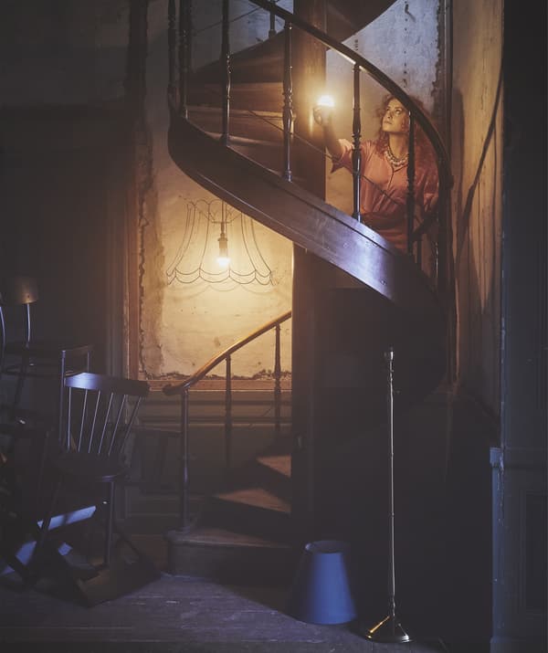 A woman lighting her way up a wooden spiral staircase in a dark room with an illuminated bulb.