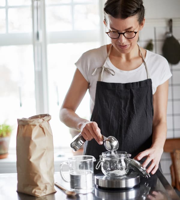 A woman measures flour into a glass jar on a kitchen scale. 