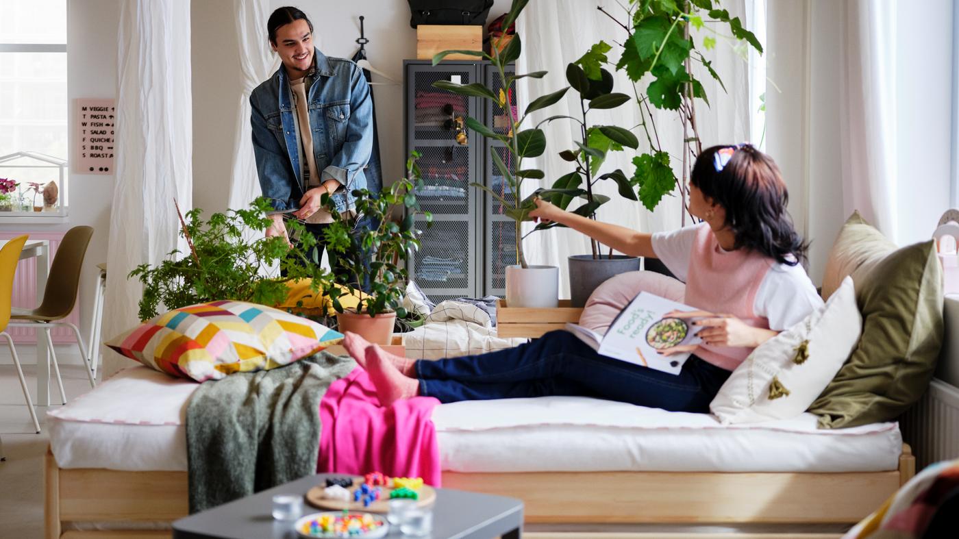 A woman on an UTÅKER bed with various colorful cushions and throws, with a man standing behind by some plants.