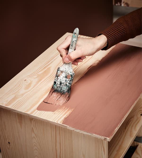 A woman paints a bedside table using a brush and brown paint to give it a new look.