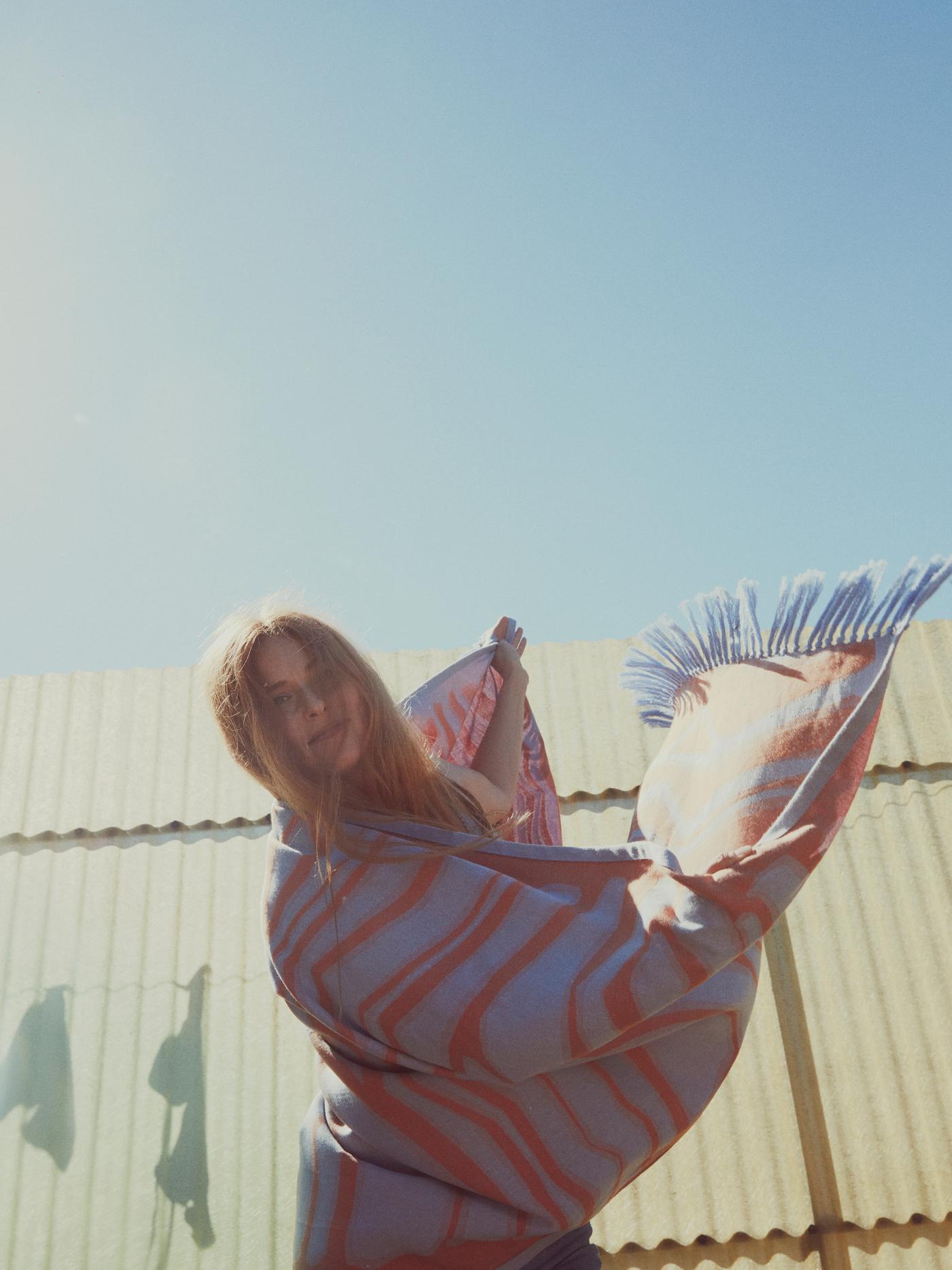 A woman playfully twirls with a blue and orange BASTUA bath sheet wrapped around her shoulders, under a clear blue sky.