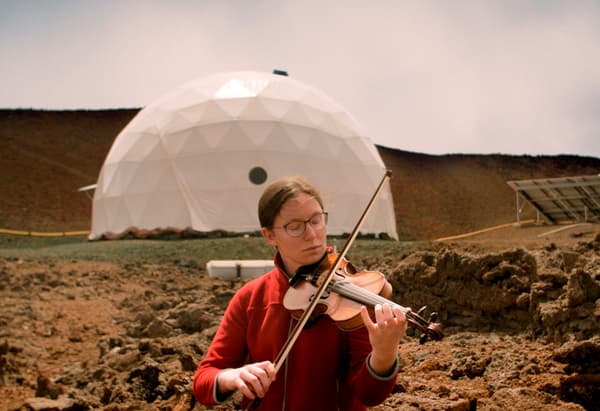 A woman playing violin in front of a white man-made structure in a barren landscape.