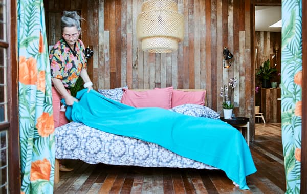 A woman puts a blue throw on a bed with purple tie-dye bedding, in a wood panelled bedroom with large woven ceiling light.