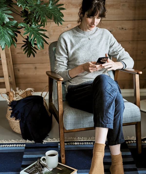 A woman scrolls through her mobile while sitting in a wood-framed armchair with grey upholstery on a blue rug.