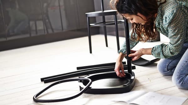 A woman sits on the floor and assembles a chair. The parts and assembly instructions lie in front of her.