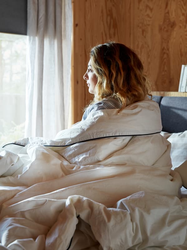 A woman sits upright in bed and looks through a sunny window, while still wrapped in a cream-colored quilt.