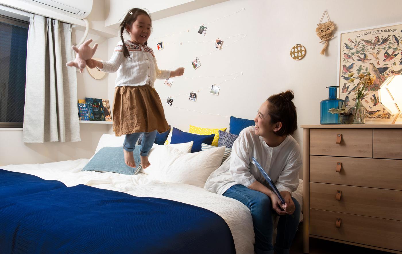 A woman sitting and a child standing on a big bed in a white bedroom with a wall-mounted bookshelf.