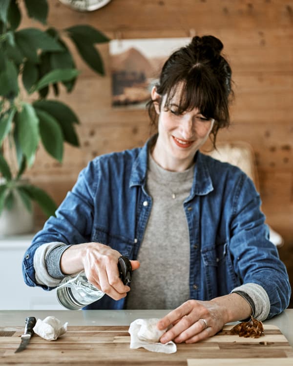 A woman sitting at a kitchen table holding a water spritzer and spraying avocado seeds wrapped in a paper kitchen towel.