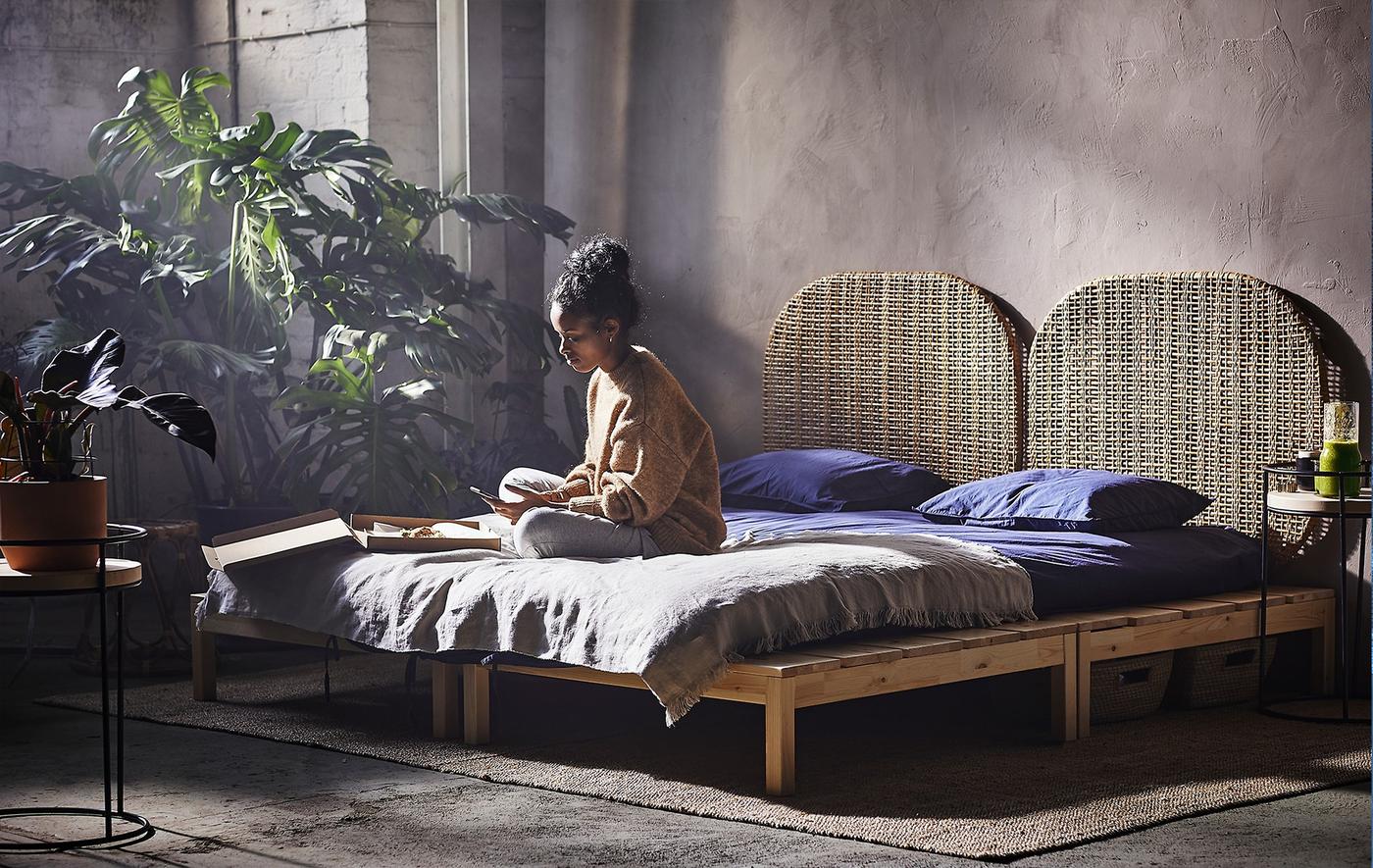 A woman sitting on a platform bed in a room with various plants for decoration.