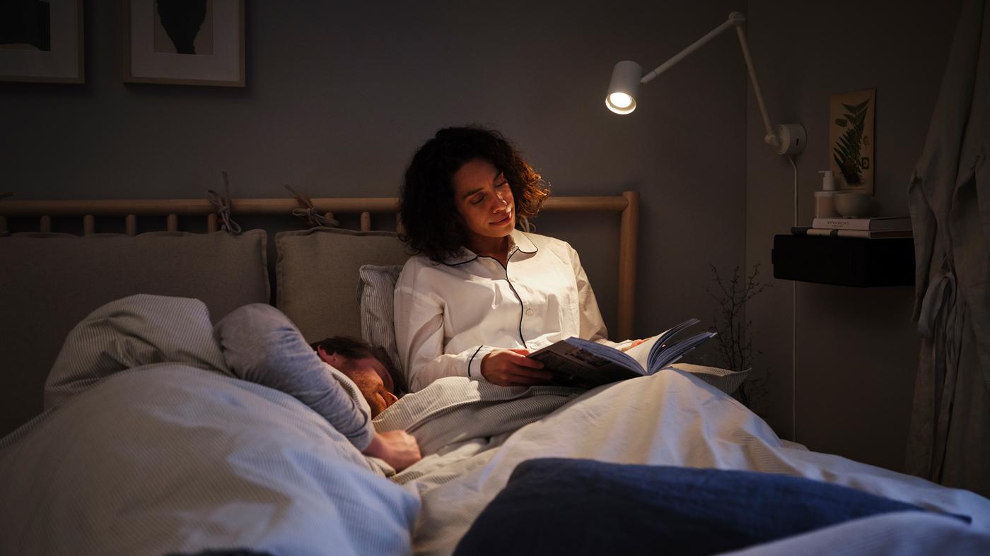 A woman sitting up in bed, reading a book under a reading lamp, with a man lying down in bed beside her.