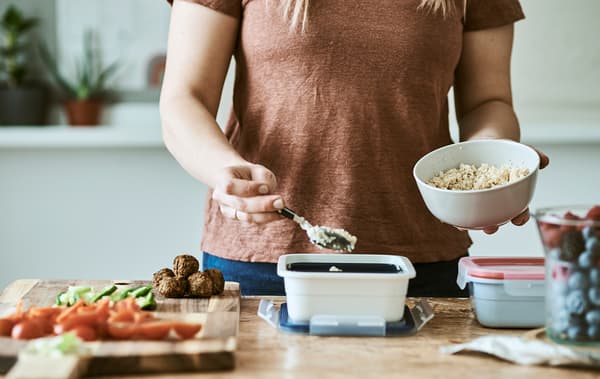 A woman spoons couscous from a white bowl into a plastic food tub. On the wood table is a chopping board with diced veg.