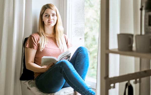 A woman wearing a T-shirt and jeans relaxes on a window seat with cushions and a white curtain, reading a book.