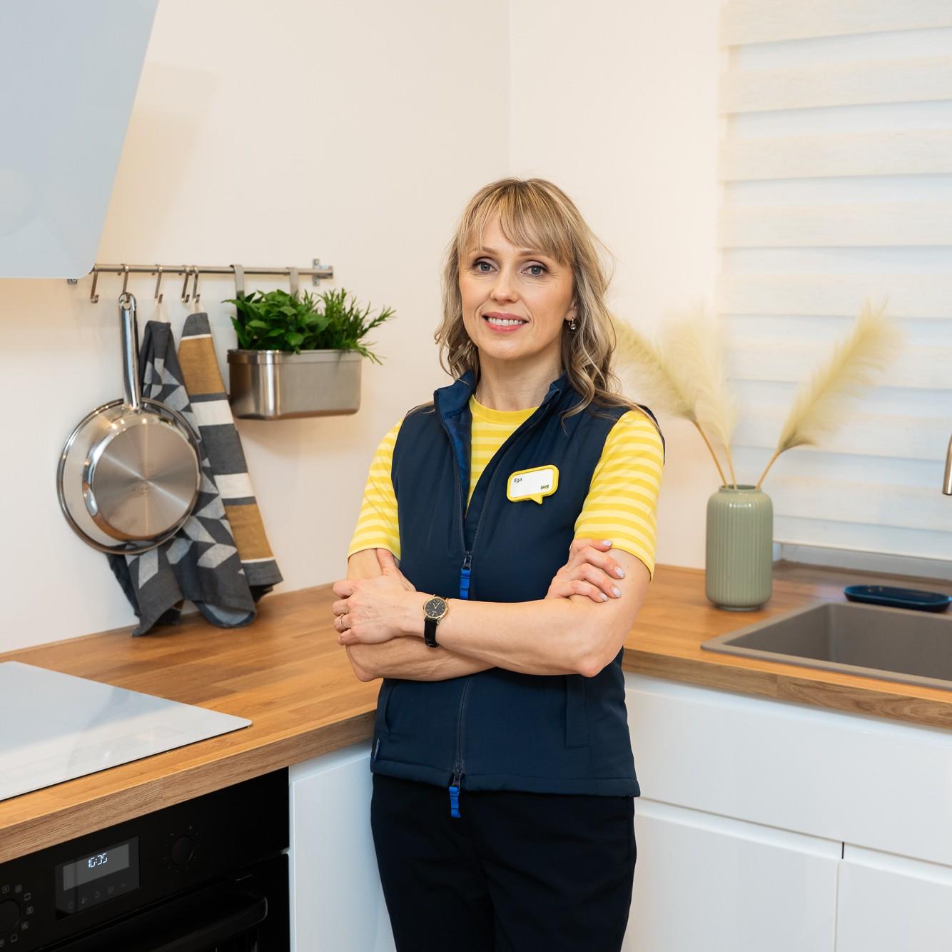 A woman wearing an IKEA uniform stands in a bright, modern kitchen with her arms crossed; a wooden worktop, white cabinets, a sink and hanging kitchen utensils are visible in the background.