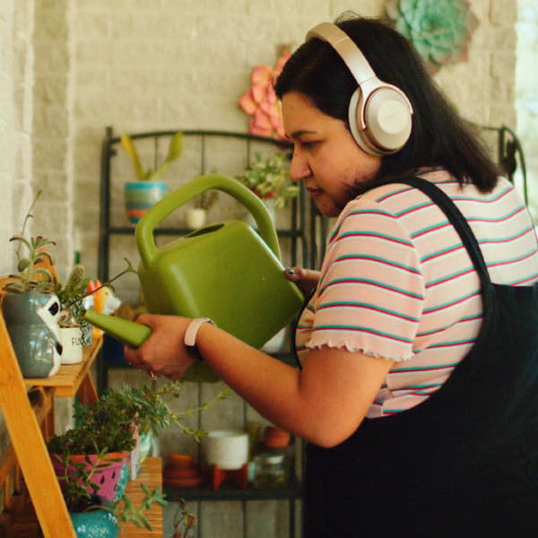 A woman wearing headphones is watering plants in multicoloured plant pots with a green watering can.