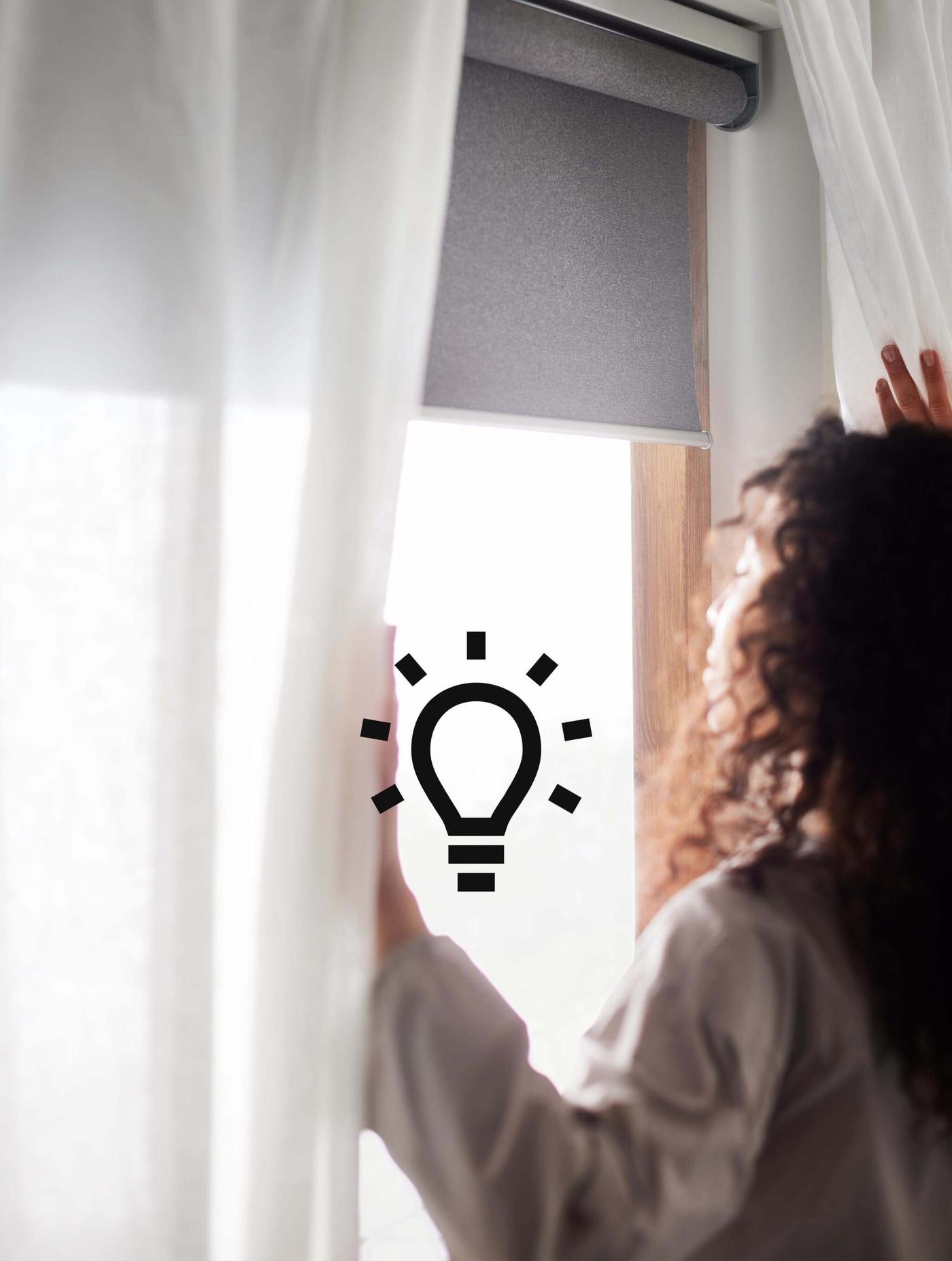 A woman with brown curly hair opening blinds and a blackout roller blind with the sun shining in.