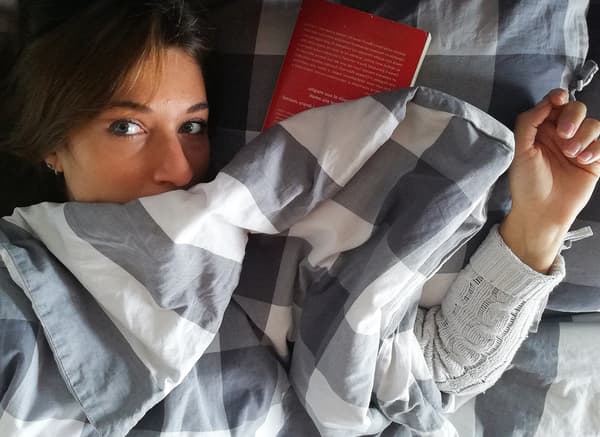 A woman with brown hair is lying underneath grey and white bedding. A red book is placed next to her head.