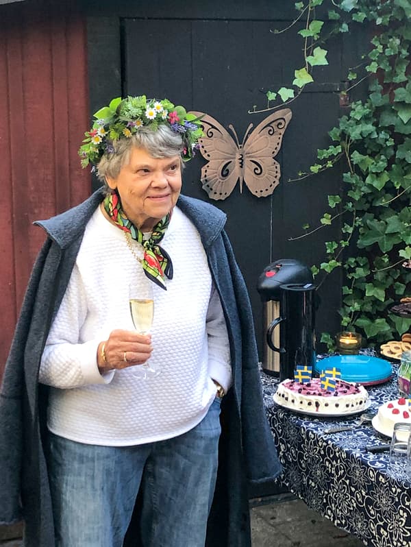 A woman with gray hair, wearing a flower crown is standing outside, holding a glass of champagne.
