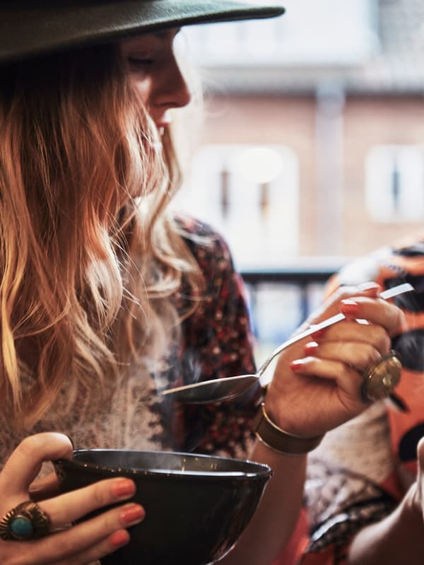 A woman with red hair is eating hot food from a bowl with a spoon. She is wearing a hat and jewellery.